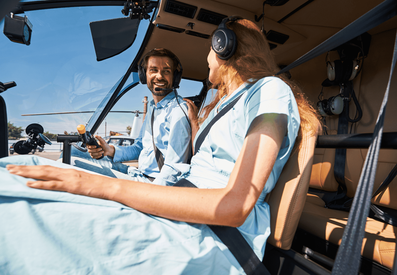 A smiling man and woman sit in the cockpit of a helicopter on a sunny day. The man is wearing a blue shirt and headset while holding the helicopter controls. The woman has long reddish hair and is also wearing a headset. They appear to be a couple on a trip. The scene has a lighthearted and happy mood. The blue sky is visible through the windshield.
