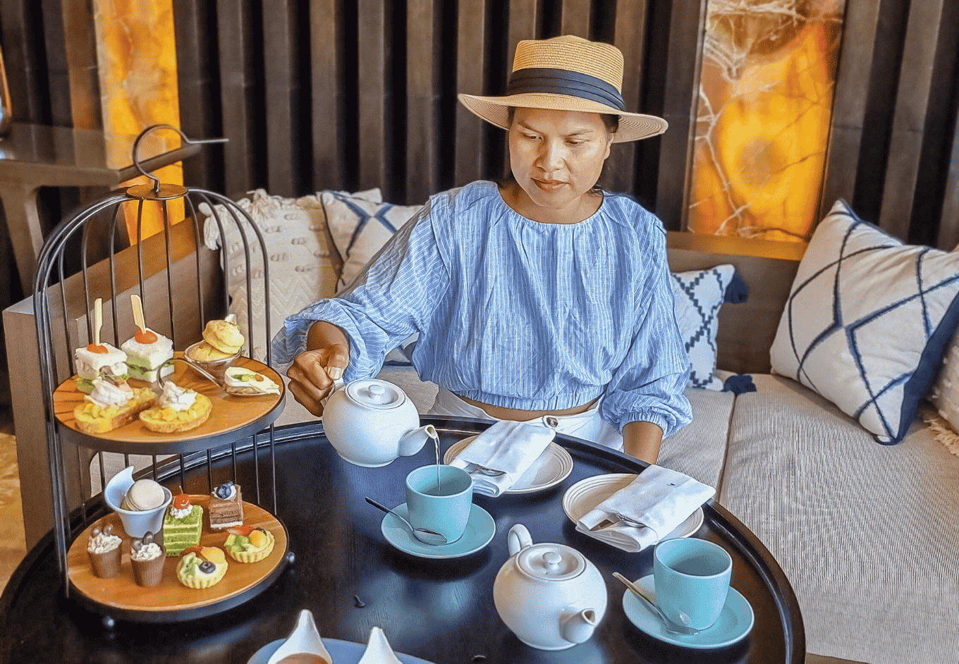 A woman in a hat pours tea from a white teapot at a round table set for afternoon tea. A tiered serving tray holds small desserts and sandwiches. The interior space has modern decor, pillows on the sofa, and a grey patterned rug. This peaceful scene could be used for projects about afternoon tea, relaxation, hotels, cafes, and travel.