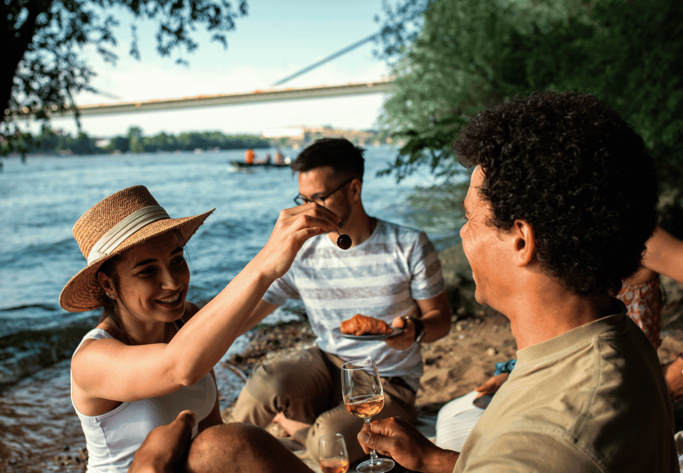 Friends gather together near the water in an urban setting. One woman with wavy brown hair and a straw hat is extending a cherry to a man with dark curly hair. Another man with glasses is holding a plate with a croissant. The group is sitting on the ground near the shore, with a bridge and water visible in the background. The scene is bathed in sunlight, with a casual, light-hearted mood. 