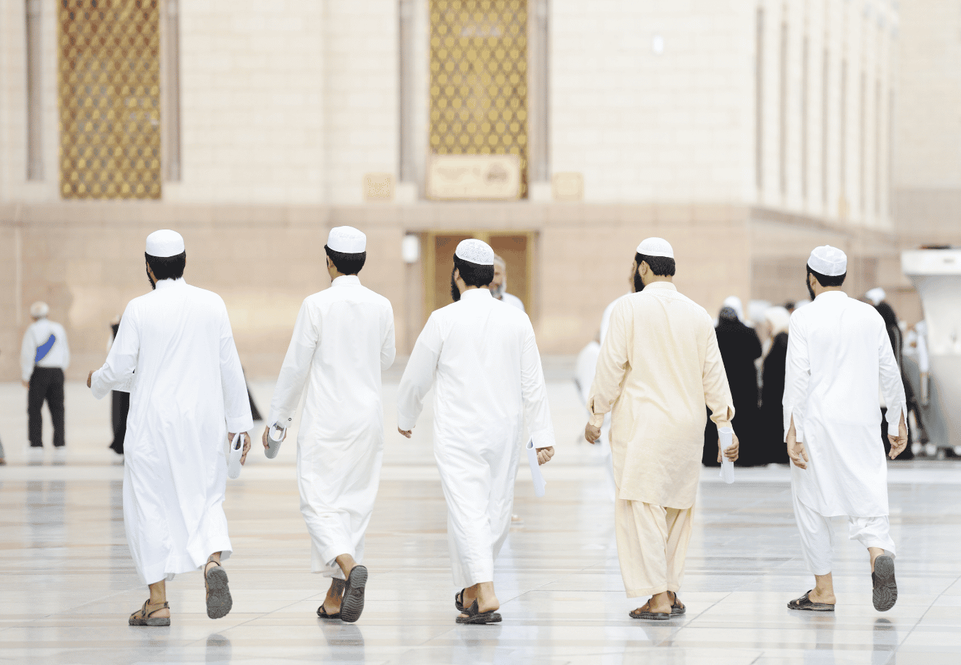 Hajj heatSeveral men are walking away from the camera, through a large open walkway. The men wear full-length traditional white and beige garments and caps. In the background is a large light colored building with gold accents. The walkway is bright and reflective.