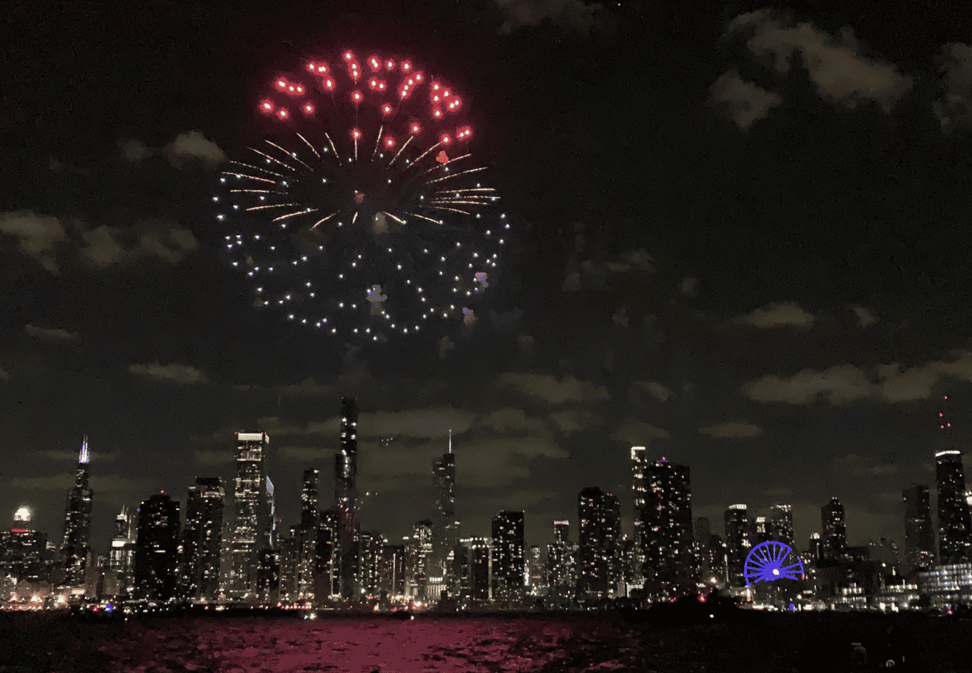Fireworks light up the night sky over an urban area. A city skyline is silhouetted against a night sky filled with clouds and red and blue fireworks. The city buildings have many illuminated windows. The water reflects red and purple lights.