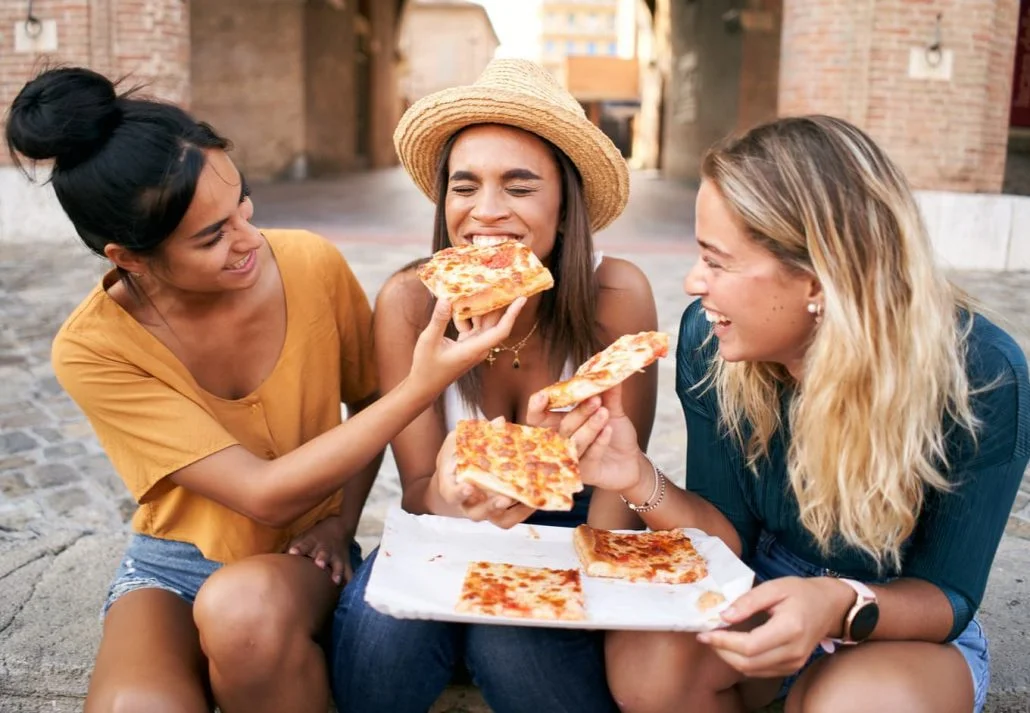 Food to try in Rome - Three young women laughing while eating pizza together in Italy.
