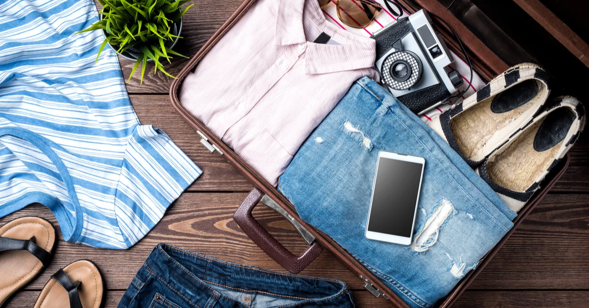 A pink shirt, jeans, shoes, a smarphone and a camera inside an open travel suitcase.