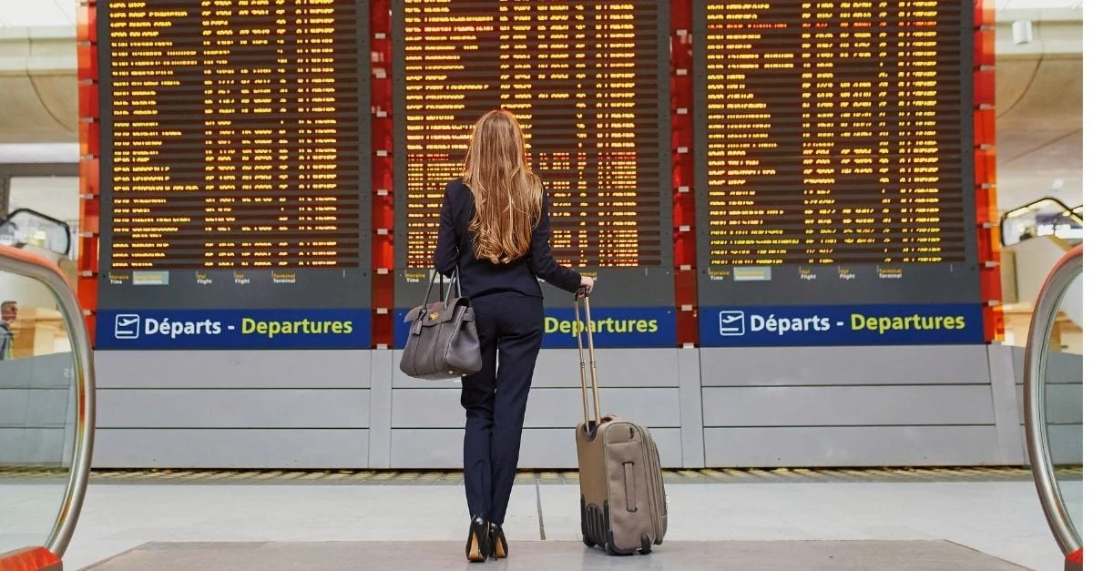 Woman checking the departures table on the airport with her carry on luggage on hand.