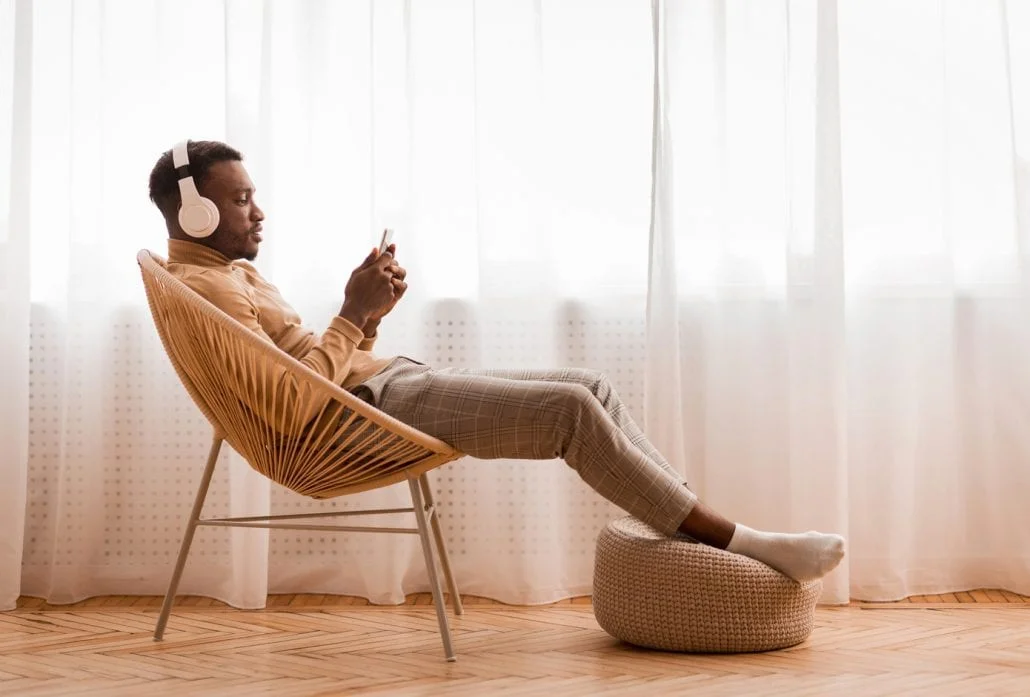 Relaxed Man Listening To Podcast Sitting On Modern Chair Against Window Indoor 