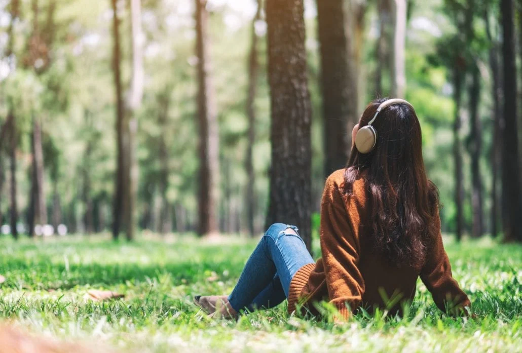 Young woman listening to music with headphones on while sitting at the grass at a park. 